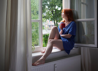 young woman with coffee mug sitting on window sill at home