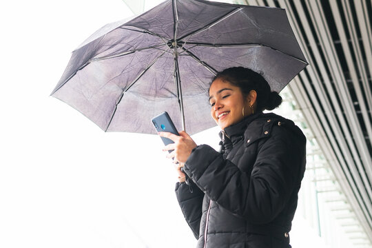 Smiling ethnic woman using smartphone under umbrella on street