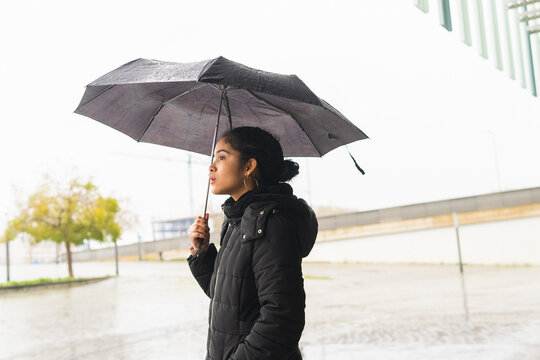 Ethnic woman with umbrella standing on street during rain