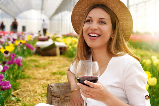Image of laughing bright woman sitting between flowers in garden outdoor bar holding wine glass on spring