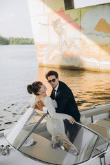 Groom in a black suit and sunglasses hugs the bride on a yacht. Wedding day