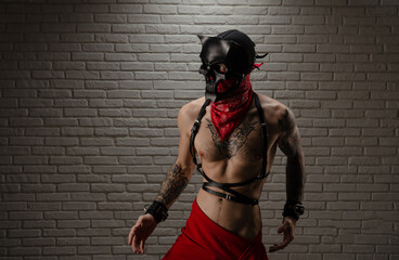 a man with a naked torso in a bdsm demon mask with tattoos and leather belts and handcuffs - obrazy, fototapety, plakaty