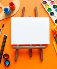 white blank sheet, wooden easel and various art supplies on orange background. copy space. place...