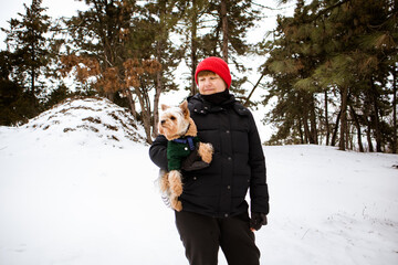 Fototapeta na wymiar A woman 30-40 years old in red cap holding hugging small Yorkshire Terrier dog walking in winter park in cold day. Pet with owner together in pine park, woods. Doggy in clothes. City people in snow.