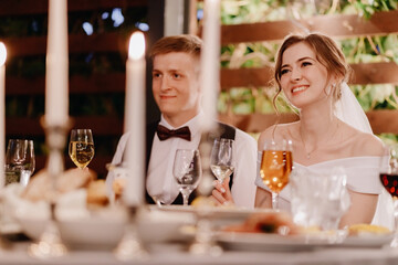 Newlyweds sit and smile with glasses at the banquet table in the evening