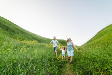 Naklejka na ściany i meble Happy young family spending time together, running on nature, on vacation, outdoors. Mom, dad, and daughter walk in the green grass. The concept of family holiday.