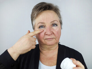 An elderly woman applies an anti-aging face cream. Cosmetology. Withering (aging) of the skin. Pigmentation. Couperose.