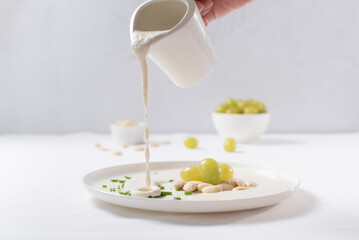 Obraz na płótnie Canvas Verter crema de almendras, sopa fría española. Ajo blanco de Málaga con almendras y uvas en un recipiente blanco sobre una mesa blanca