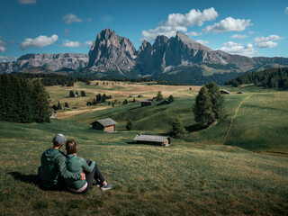 Couple sitting on meadows with wooden cabins at Alpe di Siusi during summer with view to mountains...
