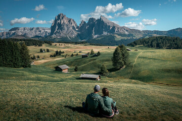 Fototapeta na wymiar Couple sitting on meadows with wooden cabins at Alpe di Siusi during summer with view to mountains of Plattkofel and Langkofel in the Dolomite Alps in South Tyrol, Italy.