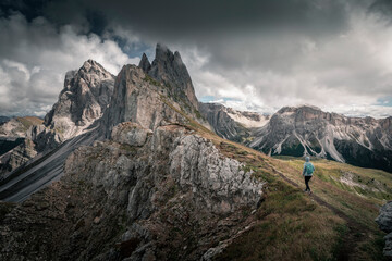 Fototapeta premium Girl hiking along cliffs of dramatic mountain peaks of Seceda with clouds in the European Dolomite Alps, meadow in the foreground, steep cliff, South Tyrol Italy.