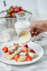 French pancakes with strawberry filling and fresh strawberries. A woman's hand holds a gravy boat and pours creamy vanilla sauce over crepes.
