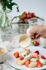 French pancakes with strawberry filling and fresh strawberries. A woman's hand holds a gravy boat and pours creamy vanilla sauce over crepes.