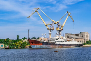 Fototapeta premium A large rusty abandoned ship against the background of two port cranes in Mykolaiv, Ukraine. Shipyard equipment on the Inhul river