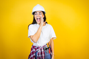 Young caucasian woman wearing hardhat and builder clothes over isolated yellow background hand on...