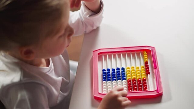 Cute little schoolgirl does sums moving plastic beads by left hand finger on colorful abacus at arithmetic lesson at home upper closeup slow motion