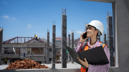 Cute-looking Asian female engineer using her phone at a construction site of houses and buildings.
woman with black hair wearing a hard hat,reflective vest ,holding files and cell phones.