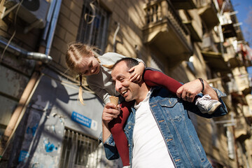 Fototapeta na wymiar Father and daughter eating ice cream outside at the street. Daughter is sitting on the fathers shoulders .