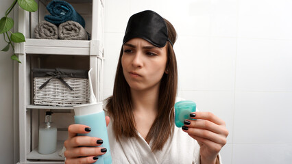 Young female with a sleeping mask on her head in a bathrobe holding dental floss in one hand and an...
