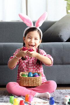 Happy adorable pink little Easter bunny girl kid with rabbit ears headband smiling and laughing after finding colorful Easter egg, egg hunt game, celebration of spring beginning.