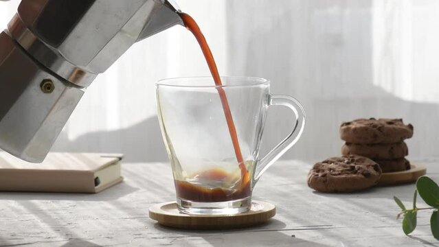 Fresh coffee is pouring from a mocha into a transparent coffee cup, next to it there are chocolate chip cookies with morning shadows and morning sunbeams through the window on a gray table background