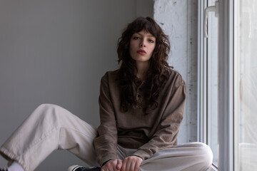 Stylish young woman with curly hair sits on the windowsill.