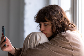 Curly girl uses a smartphone while sitting by the window.
