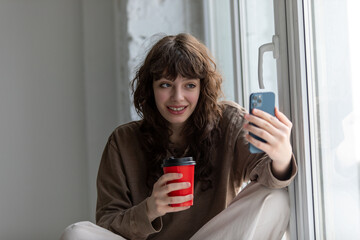 Young attractive woman with a cup of coffee in her hands makes a selfie sitting on the windowsill.