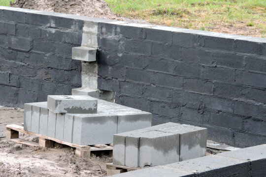 Damp proofing inside of the foundation wall with a black black asphalt-based mixture, a construction site with solid concrete blocks on pallets