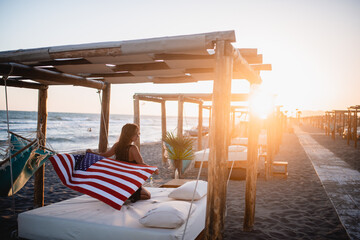 
A young girl posing with an American flag on the beach. Sunset and canopies in the background. Tan girl enjoys at the canopy