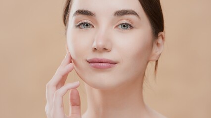 Obraz na płótnie Canvas Beauty portrait of young brown-haired European woman touches her jawline with her fingers gently and looks at camera on beige background | Skincare product commercial concept