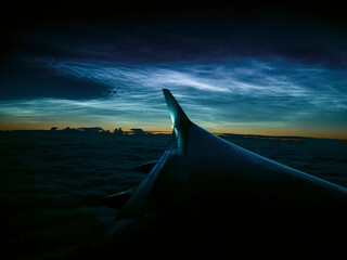 Silhouette of the left wing at cruising altitude above the clouds at night, against the backdrop of noctilucent clouds.