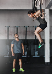 Pushing his student onto higher levels. Full length shot of a young woman working out on the gymnastics rings while her trainer looks on.