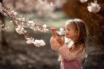 A beautiful girl with long blond hair is enjoying the flowering of almonds in the spring sunshine. Child holds twig, sniffs flowers, smiles. Good weather, a walk in the park, the onset of warm spring