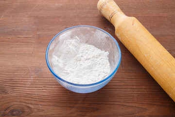 Confectionery flour in a bowl on a wooden table next to a rolling pin.