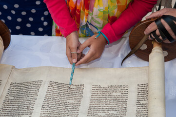 A young woman celebrating her Bat Mitzvah reads from the Torah using a colorful yad or pointer to guide her through the Hebrew text.