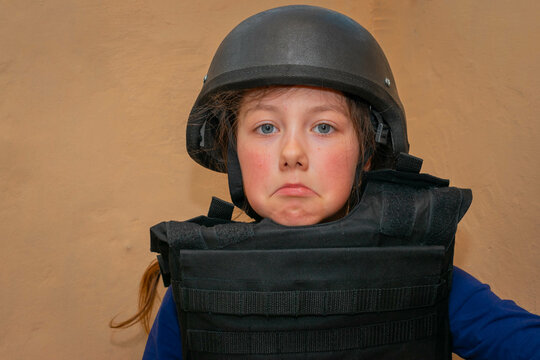 A Sad Child In A Helmet Hides From The War. A Girl In A Bulletproof Vest Sits In A Bomb Shelter