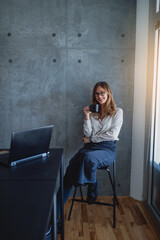 A pretty casual businesswoman with glasses sits on the chair between the desk and window in the corner of the chancellery. The gray wall is behind her. She holds a mug of tea. Posing looks at the came