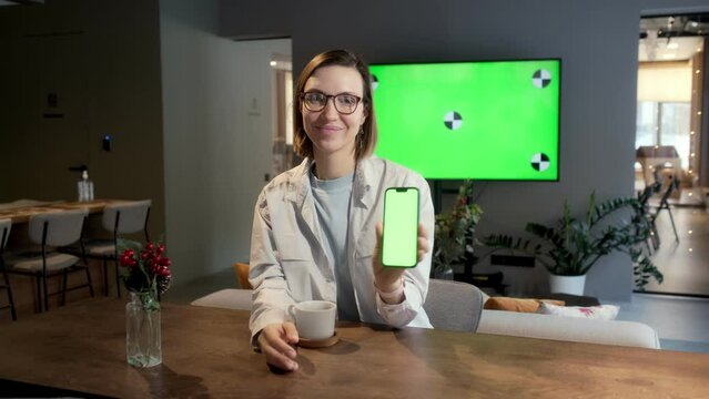 A beautiful girl with glasses sits at a table in the living room. In the background is a green screen TV. The girl raises the phone with a green screen and smiles.