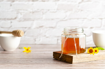 Honey in a jar on a piece of wood, a honey dripper, white bowls and some marigolds, on a wooden table and a white wall. 