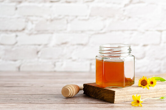 Honey in a jar on a piece of wood, a honey dripper and some marigolds, on a wooden table and a white wall. 
