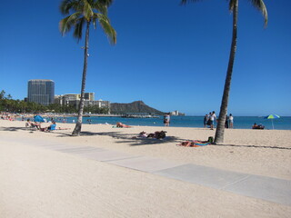 View of Kahanamoku Beach, Waikiki. View of the horizon with palm trees, Diamond Head mountain summit, clear blue sky, the skyline, and a walkaway across the beige sand. Oahu, Hawaii.