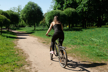 A white-skinned girl rides a bicycle through a summer park. View from the back