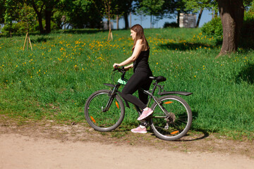 A girl on a bicycle on the background of a green park. A young brunette in a sporty black uniform on a bike ride
