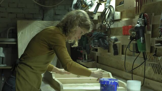 A woman paints a wooden part of a wooden product with a brush. Business idea for a home workshop