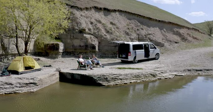 view from a drone as a guy and a girl are sitting on chairs in a campsite on the lake shore against a rock near a tent and a car. tourists rest in the mountains. overall plan