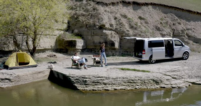 the guy and the girl are lying on a chair on the river bank near the capping machine and the tent, the girl takes a banana, puts it up and gives it to the guy. motor home, vacation in the mountains