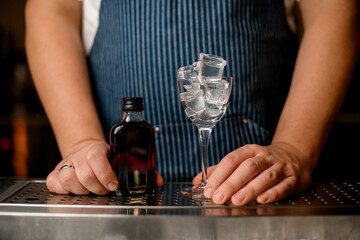 Hands of bartender holds small bottle with alcoholic drink and crystal clear empty glass