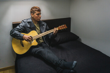 hombre tocando la guitarra sentado sobre la cama, chico joven tocando un instrumento musical en su...