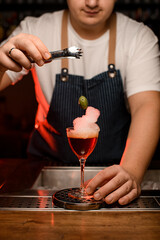 Barman using tongs to put an olive into glass filled with red foamy alcoholic drink